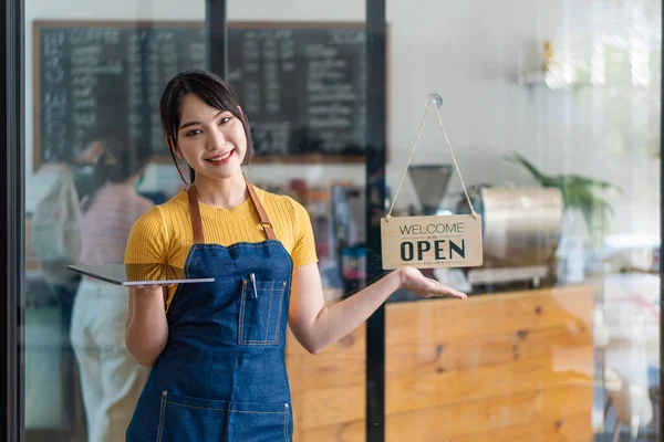 A beautiful Asian barista in an apron holds a plaque and stands in front of a cafe door with an open sign.