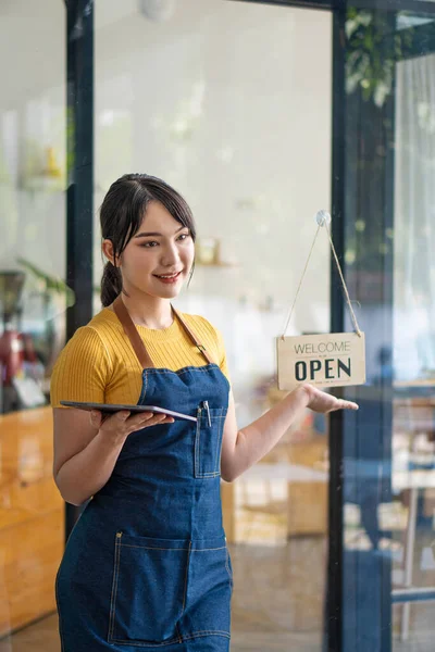 A beautiful Asian barista in an apron holds a plaque and stands in front of a cafe door with an open sign.