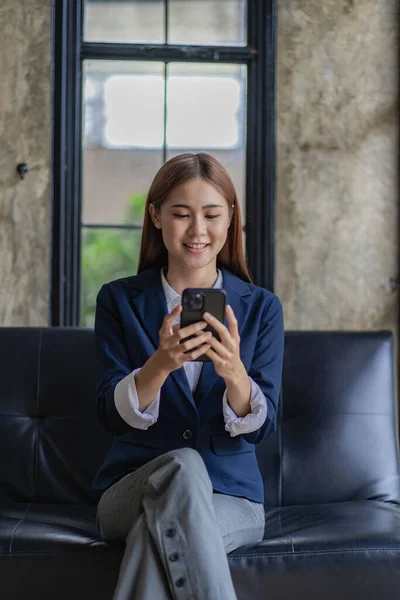 Asian woman in a suit talking on a smartphone on the sofa A cheerful business woman talks on a mobile phone via the Internet.