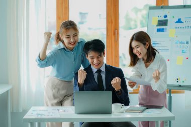 Attractive young businessman and young Asian woman celebrating successful business raising hands at their desks.