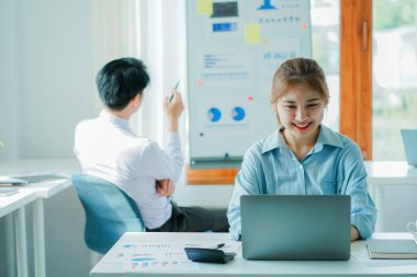 Successful Asian man, manager, businessman working with an Asian female colleague at a desk in a company office with a financial graph and laptop on the table.
