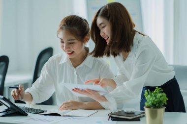 Two Asian women talk, discuss, work on the new start-up project idea. Analyze financial graphs, marketing plans and office investments.