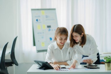 Two Asian women talk, discuss, work on the new start-up project idea. Analyze financial graphs, marketing plans and office investments.
