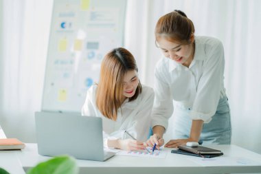 Two Asian women talk, discuss, work on the new start-up project idea. Analyze financial graphs, marketing plans and office investments.