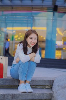 Cute Asian girl sitting in front of the mall with shopping bags. Name concept.