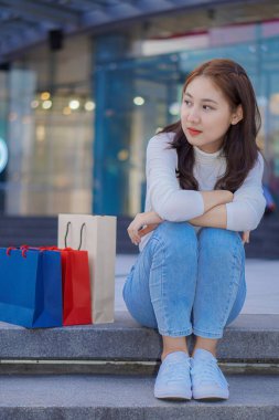 Cute Asian girl sitting in front of the mall with shopping bags. Name concept.