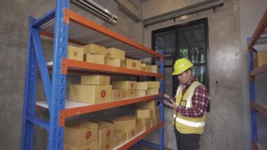 Young manager checking stock on clipboard in warehouse Side view of young Asian warehouse worker with helmet checking order details with digital tablet at inventory room.