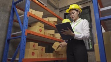 Male and female warehouse workers prepare goods for transportation.Ready stock check on clipboard in warehouse. Side view of young Asian warehouse worker with helmet checking order details
