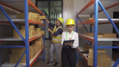 Male and female warehouse workers prepare goods for transportation.Ready stock check on clipboard in warehouse. Side view of young Asian warehouse worker with helmet checking order details