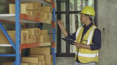 Young manager checking stock on clipboard in warehouse Side view of young Asian warehouse worker with helmet checking order details with digital tablet at inventory room.