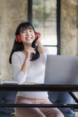 Asian woman sitting at a table and connected to a laptop She wears headphones and enjoys listening to music at work.
