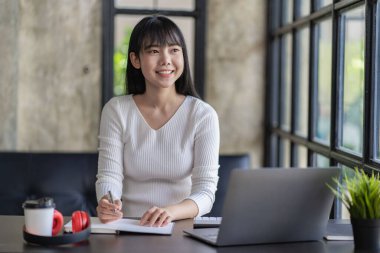Asian woman sitting at a table and connected to a laptop with headphones for working and studying online