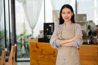 A young Asian barista holding a menu folder stands in front of a coffee shop counter, looks at the camera and smiles.