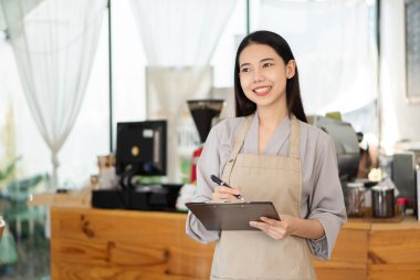 A young Asian barista holding a menu folder stands in front of a coffee shop counter, looks at the camera and smiles.