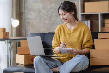 Asian woman working at home with yellow box and laptop for taking orders, sme business ideas on parcel delivery