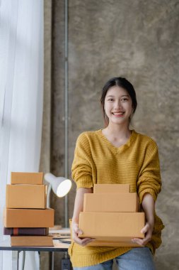 Asian woman working at home with yellow box and laptop for taking orders, sme business ideas on parcel delivery