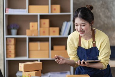 Asian woman working at home with yellow box and laptop for taking orders, sme business ideas on parcel delivery