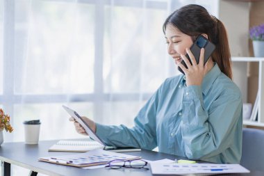 Asian woman working at home office on desk using calculator to calculate tax accounting expense report with graph document and laptop computer with smartphone.
