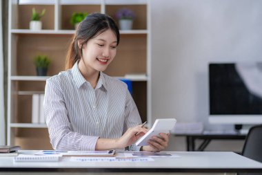 Asian woman working at home on desk using calculator to calculate tax accounting expense report with graph document and laptop computer.