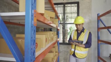 Handsome Asian employee works at a warehouse. Workers wearing high visibility clothes and helmets are checking and counting goods or boxes for delivery. Sme business idea.