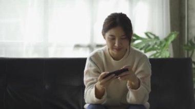 Kanepede oturan tatlı Asyalı kadın akıllı telefonuyla oyun oynuyor..