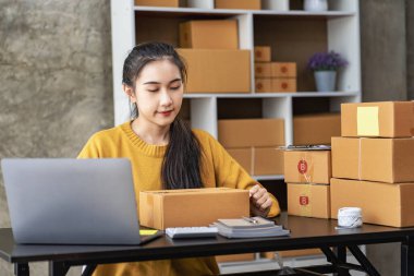 Genç Asyalı kadın evde başarılı çevrimiçi satışlar yaptı. Dizüstü bilgisayarla çalışan paketler gönderdi ve siparişleri ticari fikirlere aktardı.