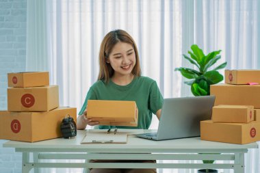 Genç Asyalı kadın evde başarılı çevrimiçi satışlar yaptı. Dizüstü bilgisayarla çalışan paketler gönderdi ve siparişleri ticari fikirlere aktardı.