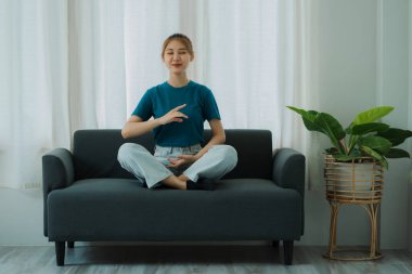 Asyalı kadın meditasyon yapıyor, kanepede oturuyor, bacak bacak bacak üstüne atıyor, çıplak ayakla katlanmış, meditasyon yapıyor, yoga yapıyor, yoga ve meditasyon gibi sakin bir odada yoga yapıyor.