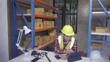 Fabrika işçileri ambara teslim edilecek paketleri denetliyor. Erkek bir depo işçisi teslimat için malları kontrol eder. Sert şapkalı bir adam tablet kullanarak kutu etiketi yazar..