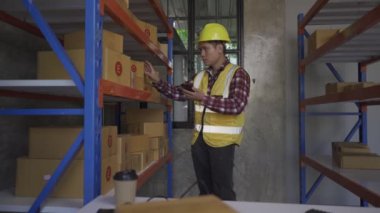 Fabrika işçileri ambara teslim edilecek paketleri denetliyor. Erkek bir depo işçisi teslimat için malları kontrol eder. Sert şapkalı bir adam tablet kullanarak kutu etiketi yazar..