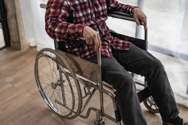 A man who can't see his face in a red checkered shirt sits in a wheelchair in the house.
