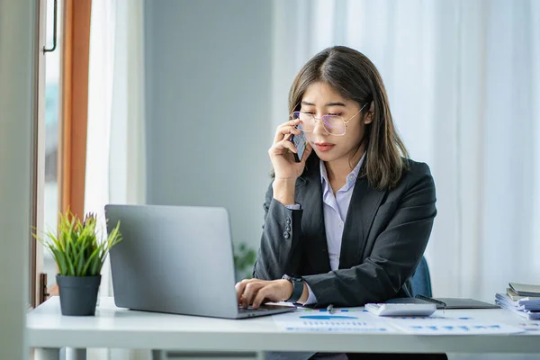 Asian businesswoman talking on the phone and doing accounting, calculating, analyzing finances with laptop in the office.