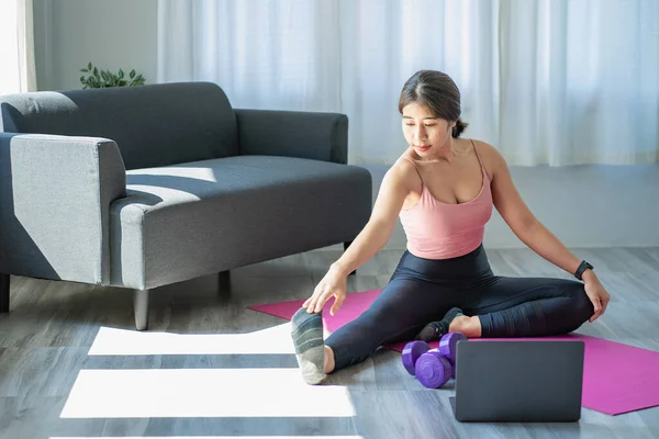 Asian woman in sportswear sitting relaxing and practicing yoga and exercising with dumbbells and laptop computer in bedroom at home fitness concept