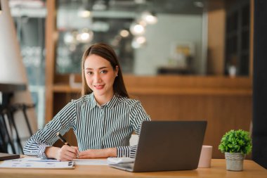 Ofiste çalışan gülümseyen iş kadını elinde para şeması belgesi ve elinde dizüstü bilgisayarla ofis muhasebe konseptinde koltukta oturuyor.