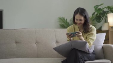 Gülümseyen Asyalı bir kadın, evinde akıllı telefondan komik içerikler seyrediyor. Neşeli bir kız, arkadaşlarıyla mesajlaşıyor. Dairede, kanepede otururken online oyun konseptinde.