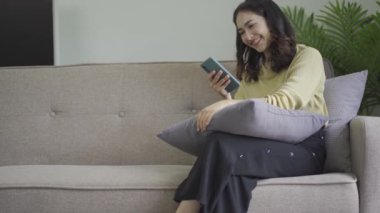 Gülümseyen Asyalı bir kadın, evinde akıllı telefondan komik içerikler seyrediyor. Neşeli bir kız, arkadaşlarıyla mesajlaşıyor. Dairede, kanepede otururken online oyun konseptinde.