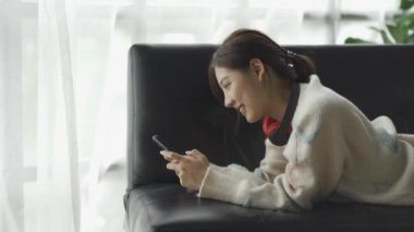 Gülümseyen Asyalı bir kadın, evinde akıllı telefondan komik içerikler seyrediyor. Neşeli bir kız, arkadaşlarıyla mesajlaşıyor. Dairede, kanepede otururken online oyun konseptinde.