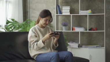 Gülümseyen Asyalı bir kadın, evinde akıllı telefondan komik içerikler seyrediyor. Neşeli bir kız, arkadaşlarıyla mesajlaşıyor. Dairede, kanepede otururken online oyun konseptinde.