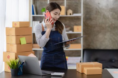Güzel Asyalı kadın evde çalışıyor. İnternette sarı kutu ve dizüstü bilgisayarla çalışıyor. Müşterilerden isim siparişleri alıyor.