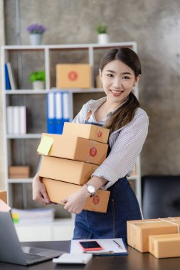Güzel Asyalı kadın evde çalışıyor. İnternette sarı kutu ve dizüstü bilgisayarla çalışıyor. Müşterilerden isim siparişleri alıyor.