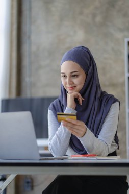 Başörtüsü takan Müslüman kadın evde kredi kartıyla internetten alışveriş yapıyor. bir dizüstü bilgisayar kullanarak ve çevrimiçi işlemler yaparak