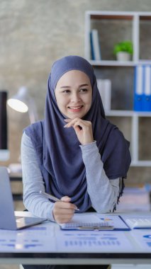 Asyalı Müslüman iş kadını dizüstü bilgisayarını kullanarak ve çizelgeler, finansal belgeler ile çalışarak, ofiste muhasebe hesaplarını analiz ederek.