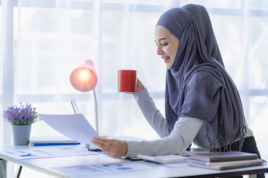 Asyalı Müslüman iş kadını dizüstü bilgisayarını kullanarak ve çizelgeler, finansal belgeler ile çalışarak, ofiste muhasebe hesaplarını analiz ederek.
