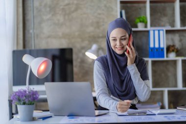 Asyalı Müslüman iş kadını dizüstü bilgisayarını kullanarak ve çizelgeler, finansal belgeler ile çalışarak, ofiste muhasebe hesaplarını analiz ederek.