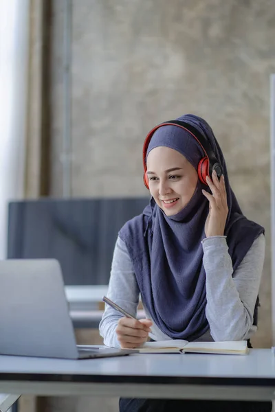 Hijab-wearing female Muslim students study online with laptop and headphones in remote online meetings.