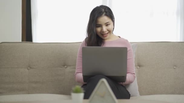 アジアの女性は 彼女の膝の上にラップトップでソファに座って 幸せな表情を示すのではなく 入力し 彼の手を上げる興奮した女性は ラップトップ上でオンラインショッピングを見て — ストック動画