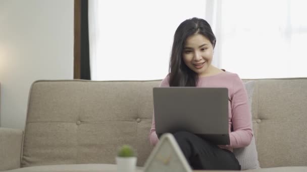 アジアの女性は 彼女の膝の上にラップトップでソファに座って 幸せな表情を示すのではなく 入力し 彼の手を上げる興奮した女性は ラップトップ上でオンラインショッピングを見て — ストック動画