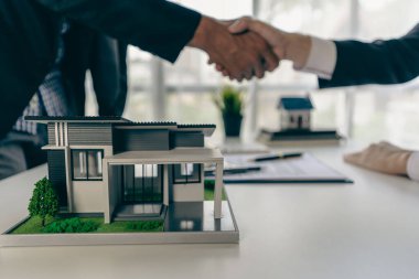 Bir emlakçı, bir ev sözleşmesiyle ilgili anlaşmayı tamamladıktan sonra bir müşteriyle tokalaşıyor. Sözleşme belgeleri ve ahşap bir masada bir ev modeli..