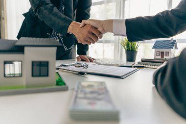 Bir emlakçı, bir ev sözleşmesiyle ilgili anlaşmayı tamamladıktan sonra bir müşteriyle tokalaşıyor. Sözleşme belgeleri ve ahşap bir masada bir ev modeli..