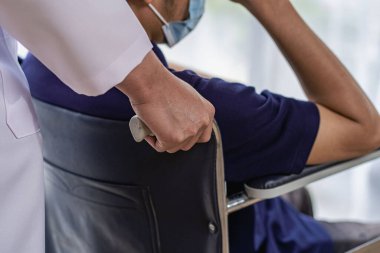 Kadın bir doktor, hastanede tekerlekli sandalyedeki bir erkek hastayı itiyor. Tekerlekli sandalyede oturan adamla hemşire. Asyalı kadınlar, beyaz erkekler.